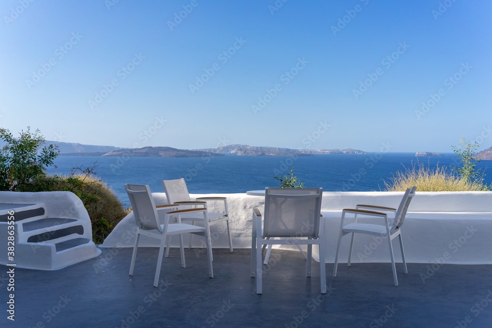 Four white chairs with white table on the white terrace with blue sea and caldera view on a sunny day, without people. Oia, Santorini island, Greece