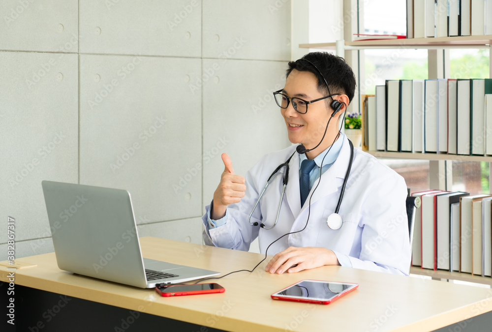 young asian doctor wearing headphone and using computer to give consult to the patient via video call in the hospital office. telemedine and healthcare concept
