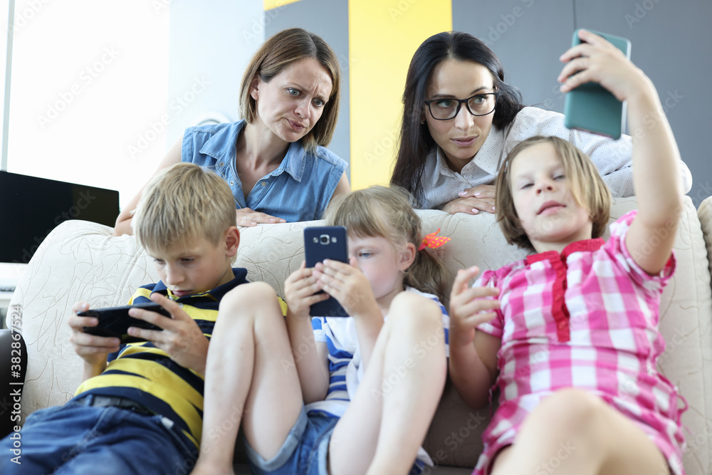 Three children sit side by side on couch and play games on phone. One woman look unhappily at daughter phone screen. Mother in glasses look surprised at girl phone. Three children sit side by side on couch and play games on phone. One woman look unhappily at daughter phone screen. Mother in glasses look surprised at girl phone.
