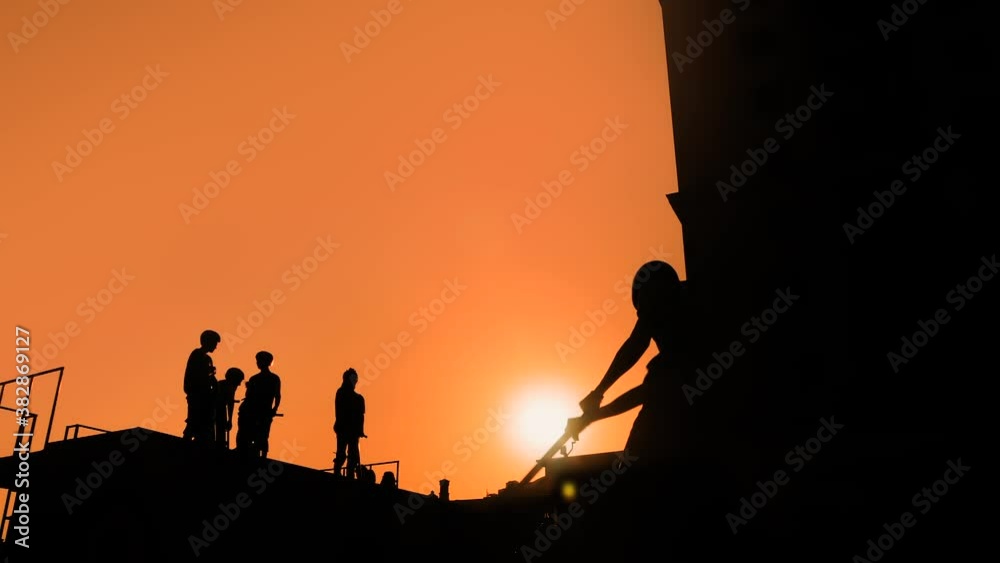 Slow motion, sun lens flare: unrecognizable teenage boy silhouette showing high jump tricks on scooter against orange sunset sky at skatepark. Sport, extreme, freestyle, outdoor activity concept