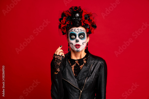 Scary young creepy lady calavera. wears artistic make-up for the feast of all the dead. Has frightened expression, wears black leather jacket and lace gloves, dressed as skeleton isolated in red.