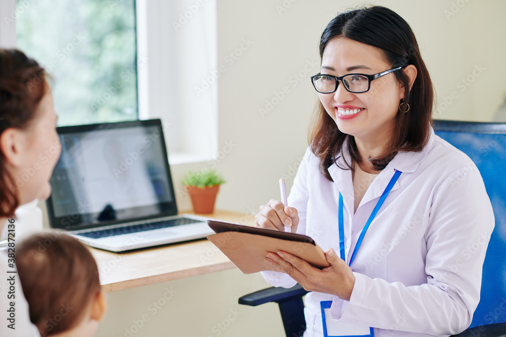 Pretty smiling pediatrician taking notes when talking to mother of little baby girl Pretty smiling pediatrician taking notes when talking to mother of little baby girl