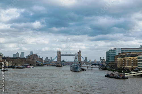 Along the Thames river in a cloudy sky you can see different types of boats such as the HMS Belfast and the London Bridge. Photograph taken in London, England, United Kingdom.