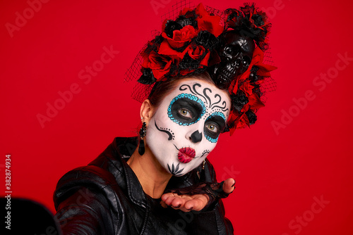 Closeup photo of funny folklore witch creature character death day facial creepy makeup masquerade. Sugar skull girl take selfies wear floral headwear amd lether jaket isolated on red background
