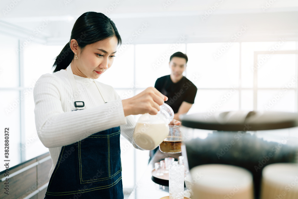 Fototapeta premium A female barista student attentively measured and added milk in the coffee while the teacher watching her in the coffee shop. Coffee making classes for entrepreneurs to start small businesses.