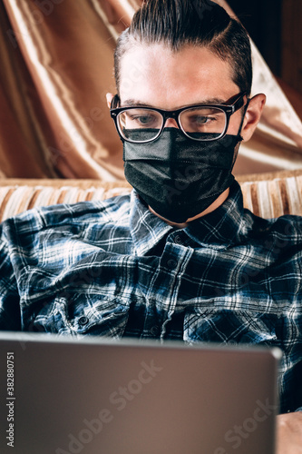 Remote operation during the quarantine, to print the text and conduct important business. A handsome young freelancer with diopter glasses and a black protective mask works on a silver laptop at home.