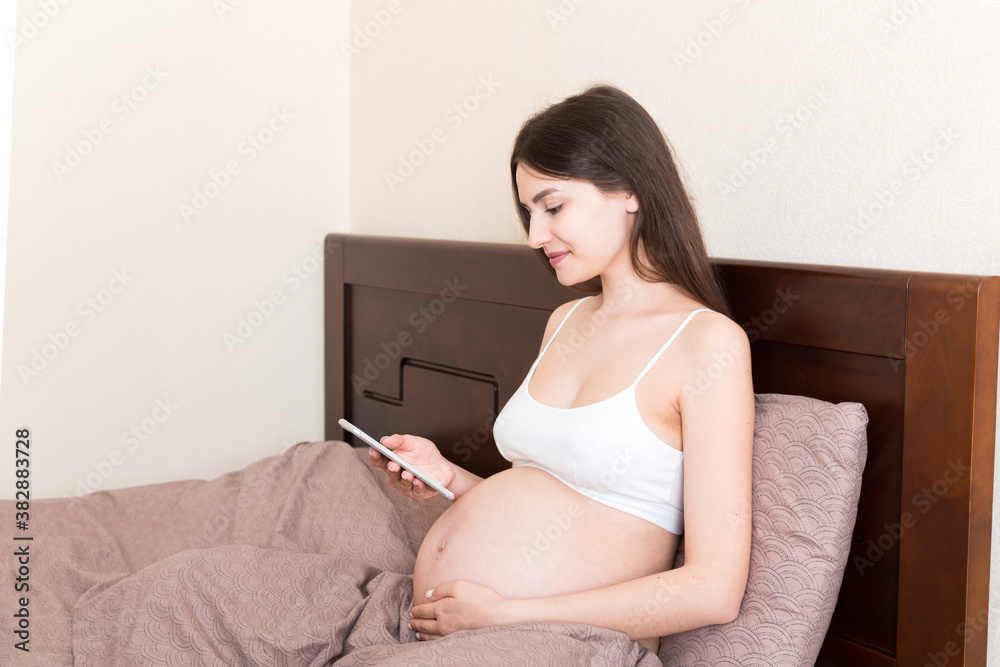 Caucasian pregnant girl using cellphone, resting on bed in home Caucasian pregnant girl using cellphone, resting on bed in home