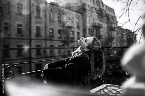 Black and white photo of stylish woman posing on the balcony in the city. Spring autumn fashion concept. Fashionable woman resting on terrace.