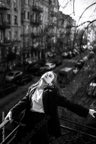 Black and white photo of stylish woman posing on the balcony in the city. Spring autumn fashion concept. Fashionable woman resting on terrace.