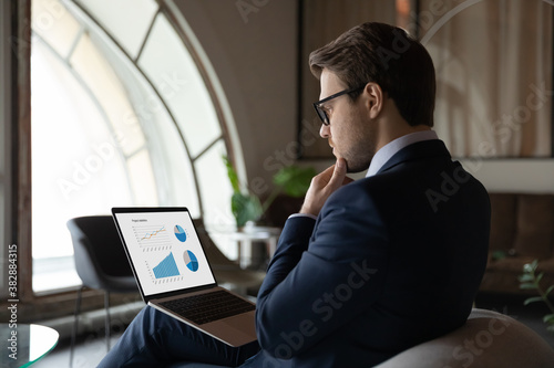Obraz na plátně Side back view focused skilled young 30s male marketing specialist in suit analyzing graphs and charts in electronic research report on computer, developing sales growth strategy alone in office
