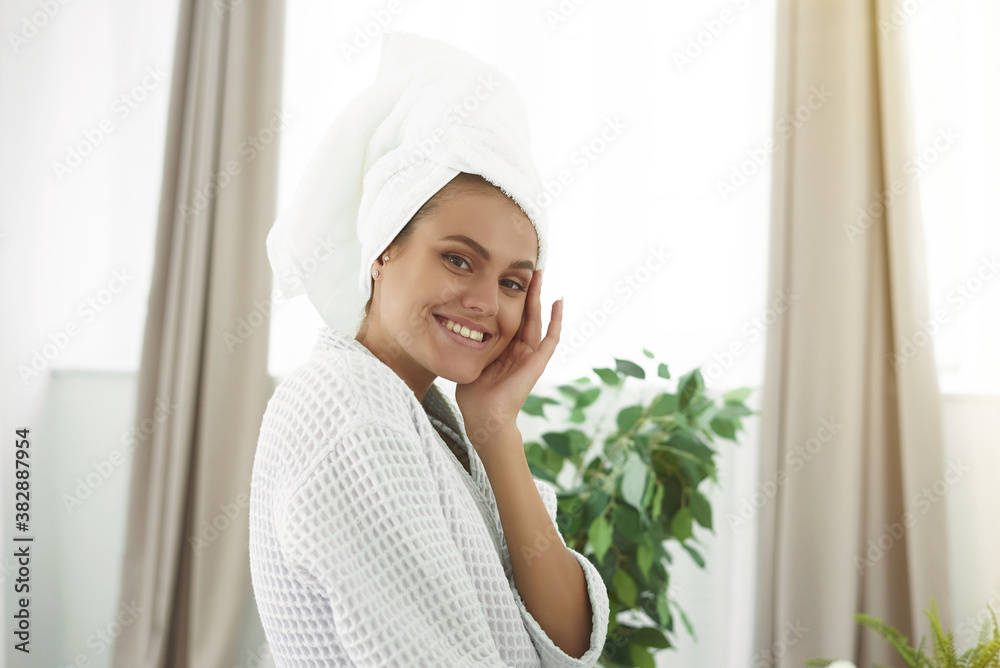 Beautiful young woman in bathrobe and towel on head smiling at camera in bathroom Beautiful young woman in bathrobe and towel on head smiling at camera in bathroom