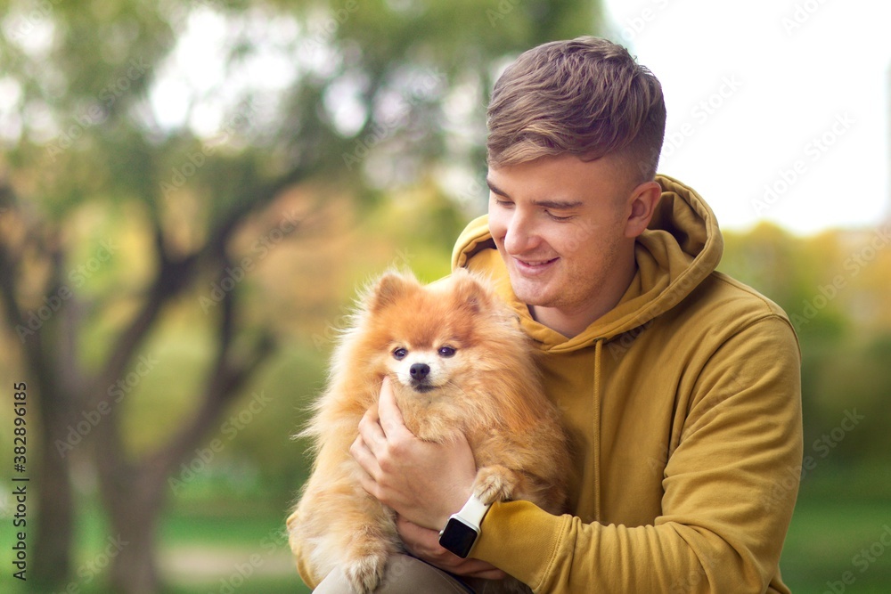 Young happy handsome man walking in autumn park, holding on hands, hugging his cute little Pomeranian spitz dog outdoors and smiling Young happy handsome man walking in autumn park, holding on hands, hugging his cute little Pomeranian spitz dog outdoors and smiling
