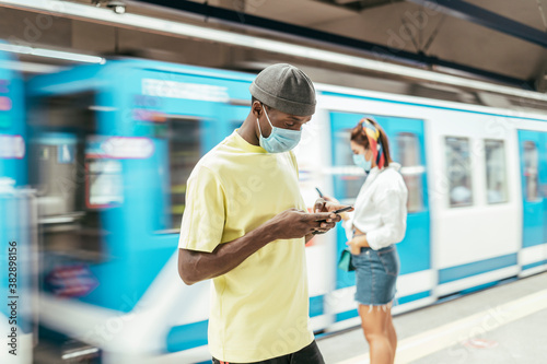 Black man with protective mask on face using smart phone while standing against subway in motion