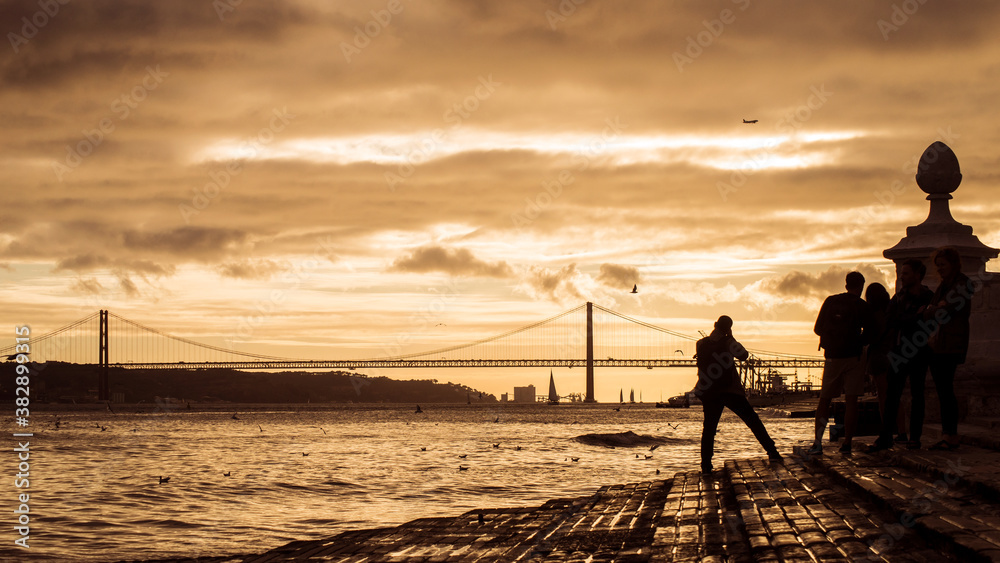 silhouette of tourist taking a picture of the famous 25th April suspension bridge on sunset background