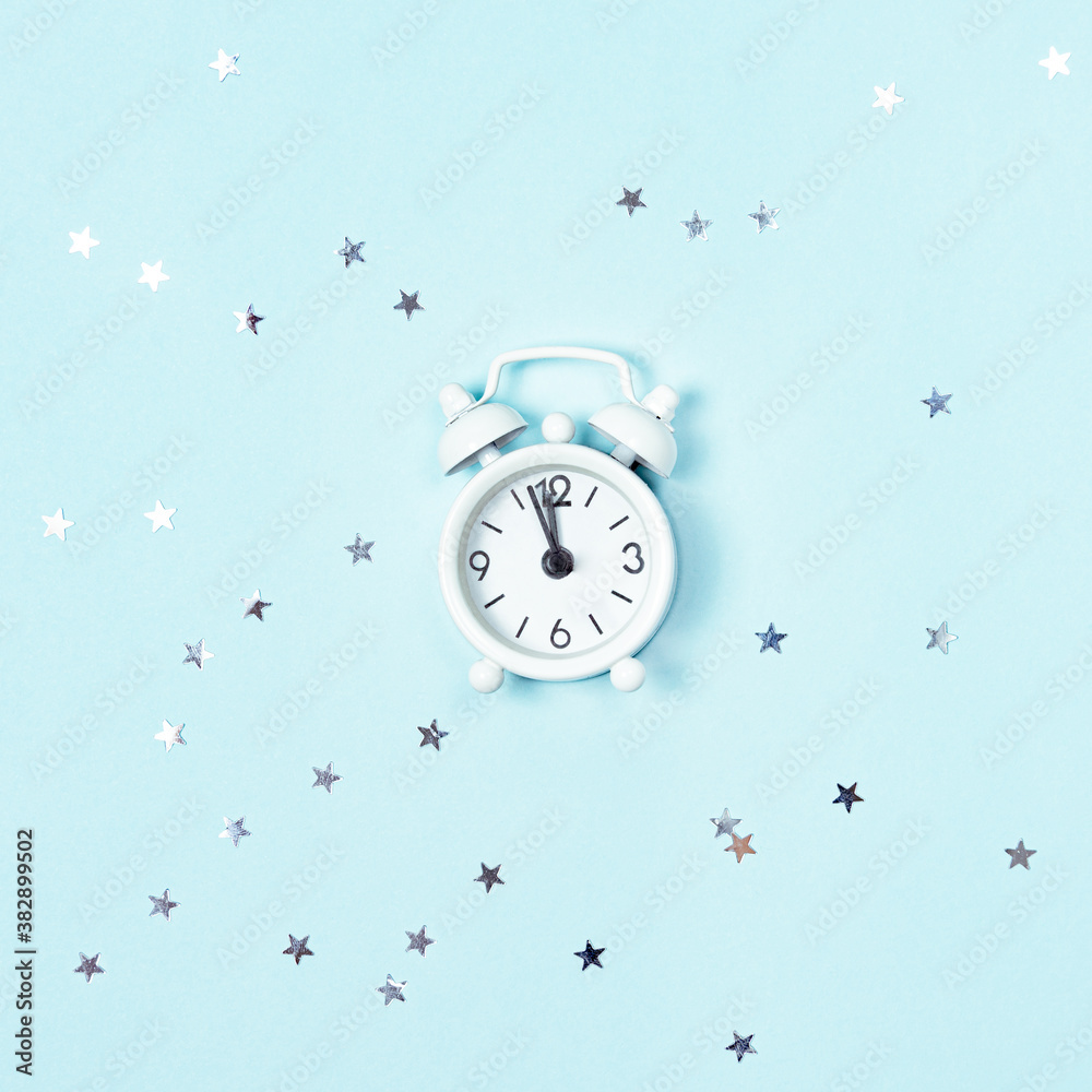 White vintage style alarm clock showing five minutes left until midnight over blue background with silver stars sequins. New Year advent.