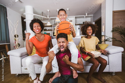 Happy African American family of three watching tv and cheering basketball games on sofa at home. © Mediteraneo