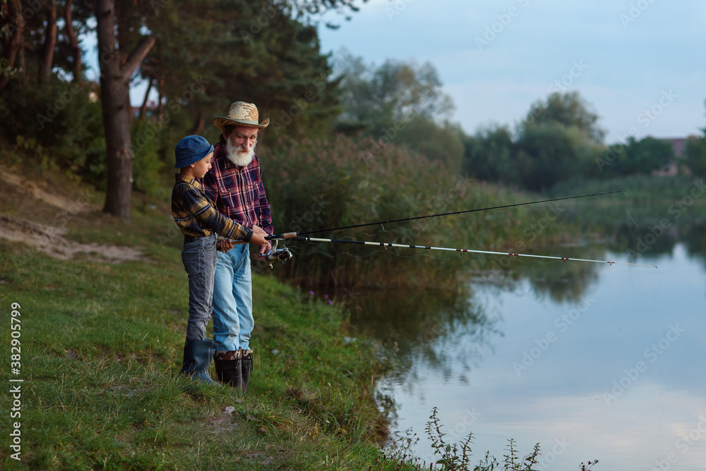Stylish experienced bearded grandpa in straw hat fishing together with his curious grandchild on the lake at sunset. Stylish experienced bearded grandpa in straw hat fishing together with his curious grandchild on the lake at sunset.