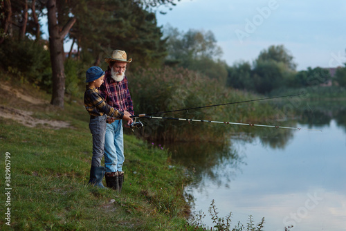 Stylish experienced bearded grandpa in straw hat fishing together with his curious grandchild on the lake at sunset.