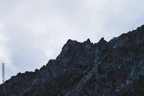 Dramatic bleak fog among giant rocky mountains. Ghostly atmospheric view to big cliff in cloudy sky. Low clouds and beautiful rockies. Minimalist scenery in mysterious place at early foggy morning.