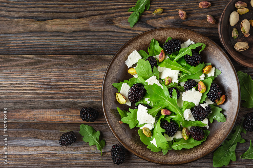 Fresh salad with arugula, feta cheese, blackberries and pistachios in a plate on rustic wooden background