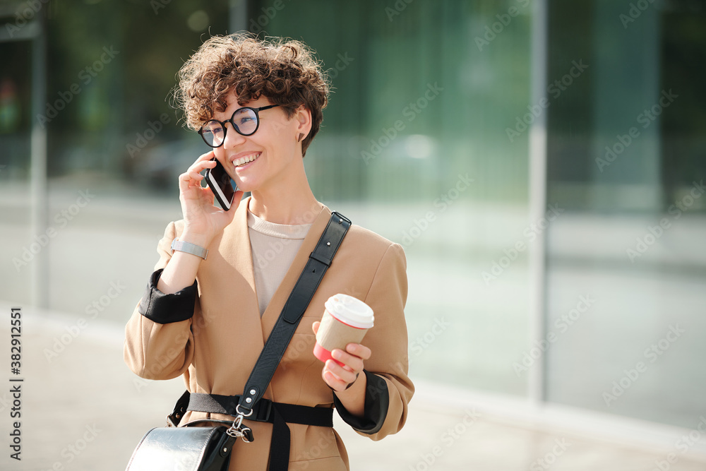 Fototapeta premium Young cheerful businesswoman with drink in hand and smartphone by ear