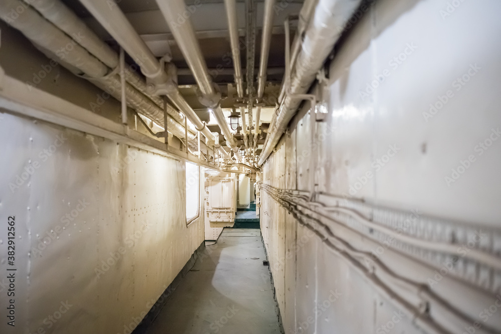 Old metal hallway or tunnel inside of an old warship. Visible piping and electrical connections on the walls and ceiling.