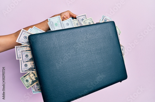 Hand of hispanic man holding briefcase with dollars over isolated pink background. photo