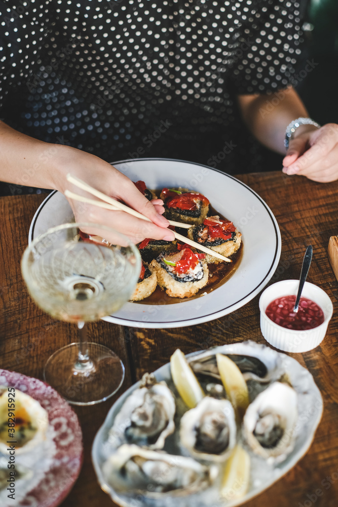 Girl eating baked susi maki with chopsticks over fresh oysters and glass of champagne on table at Japanese restaurant, top view. Japanese cuisine, fast food, seafood, fine dining concept
