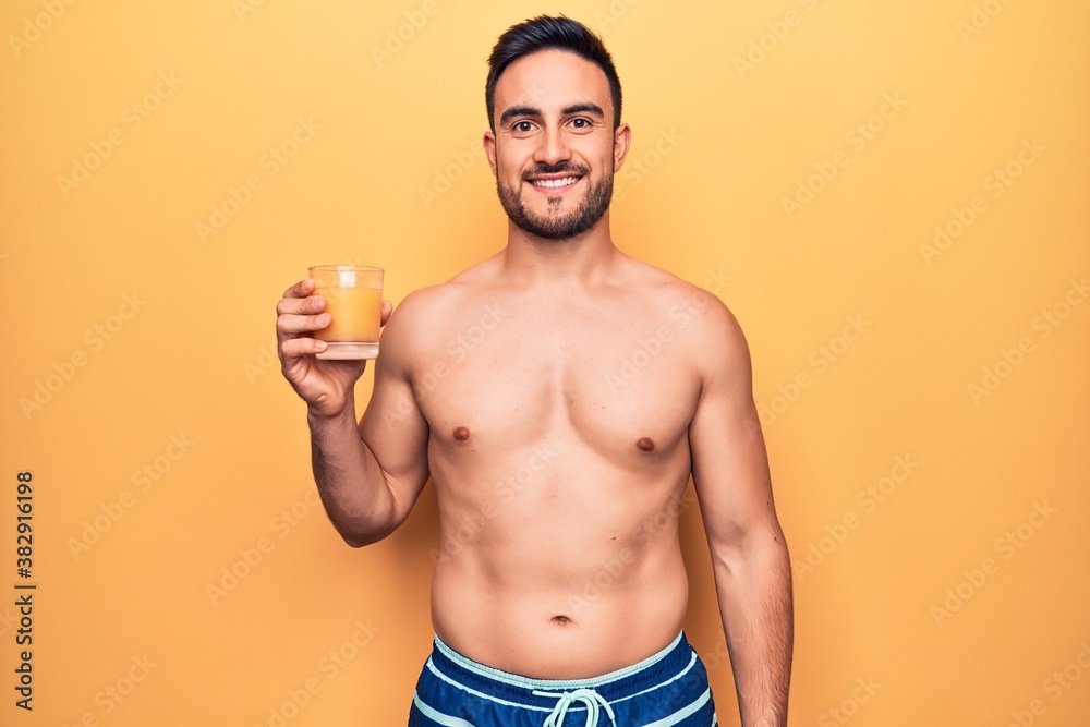 Young handsome man with beard on vacation wearing swimwear drinking glass of orange juice looking positive and happy standing and smiling with a confident smile showing teeth