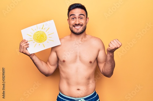 Young handsome man with beard on vacation wearing swimwear holding paper with sun draw screaming proud, celebrating victory and success very excited with raised arm