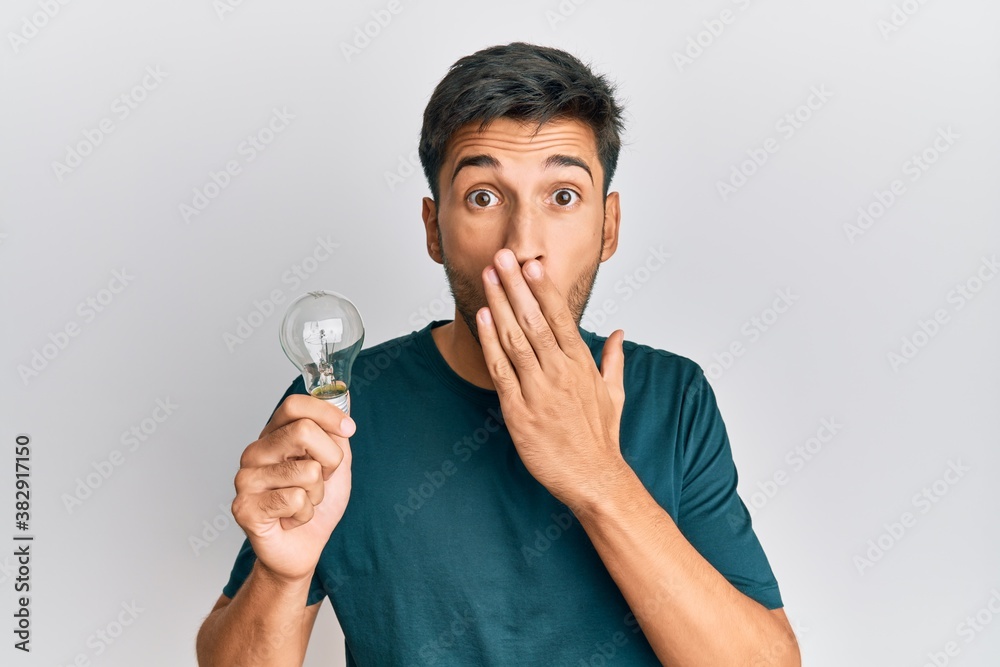 Young handsome man holding lightbulb for inspiration and idea covering mouth with hand, shocked and afraid for mistake. surprised expression Young handsome man holding lightbulb for inspiration and idea covering mouth with hand, shocked and afraid for mistake. surprised expression