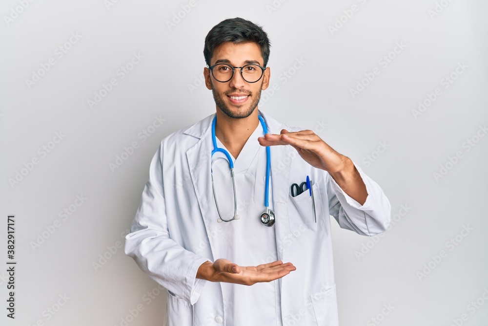 Young handsome man wearing doctor uniform and stethoscope gesturing with hands showing big and large size sign, measure symbol. smiling looking at the camera. measuring concept. Young handsome man wearing doctor uniform and stethoscope gesturing with hands showing big and large size sign, measure symbol. smiling looking at the camera. measuring concept.