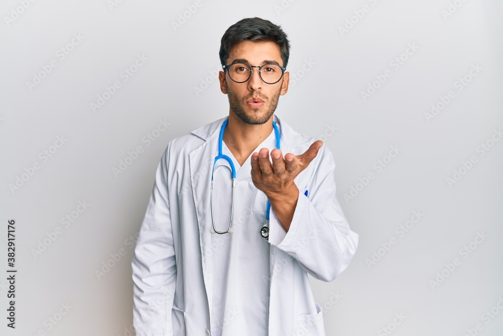 Young handsome man wearing doctor uniform and stethoscope looking at the camera blowing a kiss with hand on air being lovely and sexy. love expression. Young handsome man wearing doctor uniform and stethoscope looking at the camera blowing a kiss with hand on air being lovely and sexy. love expression.