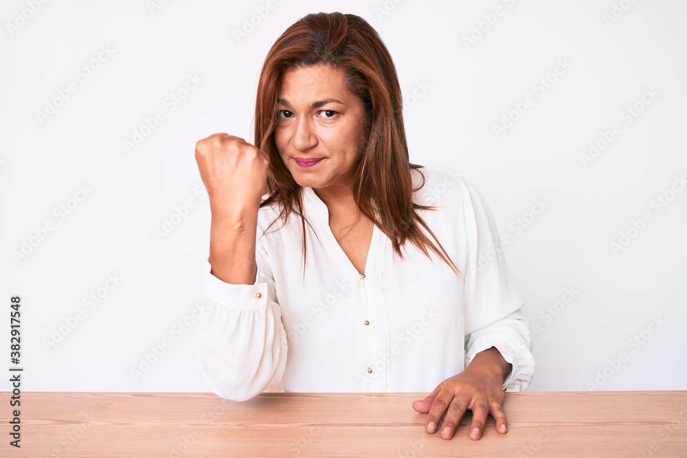 Middle age brunette hispanic business woman wearing casual white shirt sitting on the table angry and mad raising fist frustrated and furious while shouting with anger. rage and aggressive concept. Middle age brunette hispanic business woman wearing casual white shirt sitting on the table angry and mad raising fist frustrated and furious while shouting with anger. rage and aggressive concept.