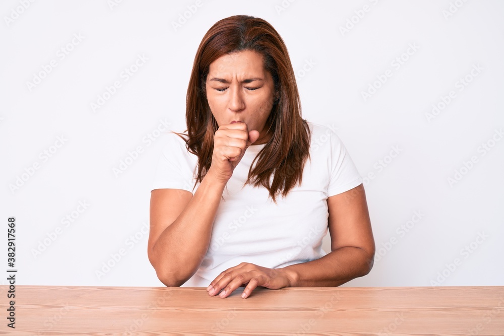Middle age brunette hispanic woman wearing casual white tshirt sitting on the table feeling unwell and coughing as symptom for cold or bronchitis. health care concept. Middle age brunette hispanic woman wearing casual white tshirt sitting on the table feeling unwell and coughing as symptom for cold or bronchitis. health care concept.