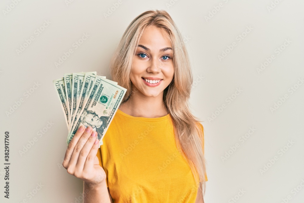 Beautiful caucasian blonde girl holding 20 dollars banknotes looking positive and happy standing and smiling with a confident smile showing teeth Beautiful caucasian blonde girl holding 20 dollars banknotes looking positive and happy standing and smiling with a confident smile showing teeth