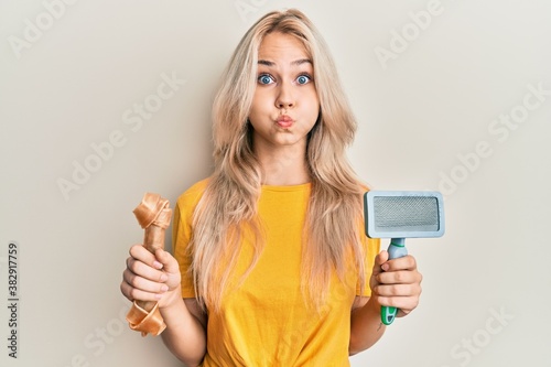 Beautiful caucasian blonde girl holding dog accesories puffing cheeks with funny face. mouth inflated with air, catching air. photo