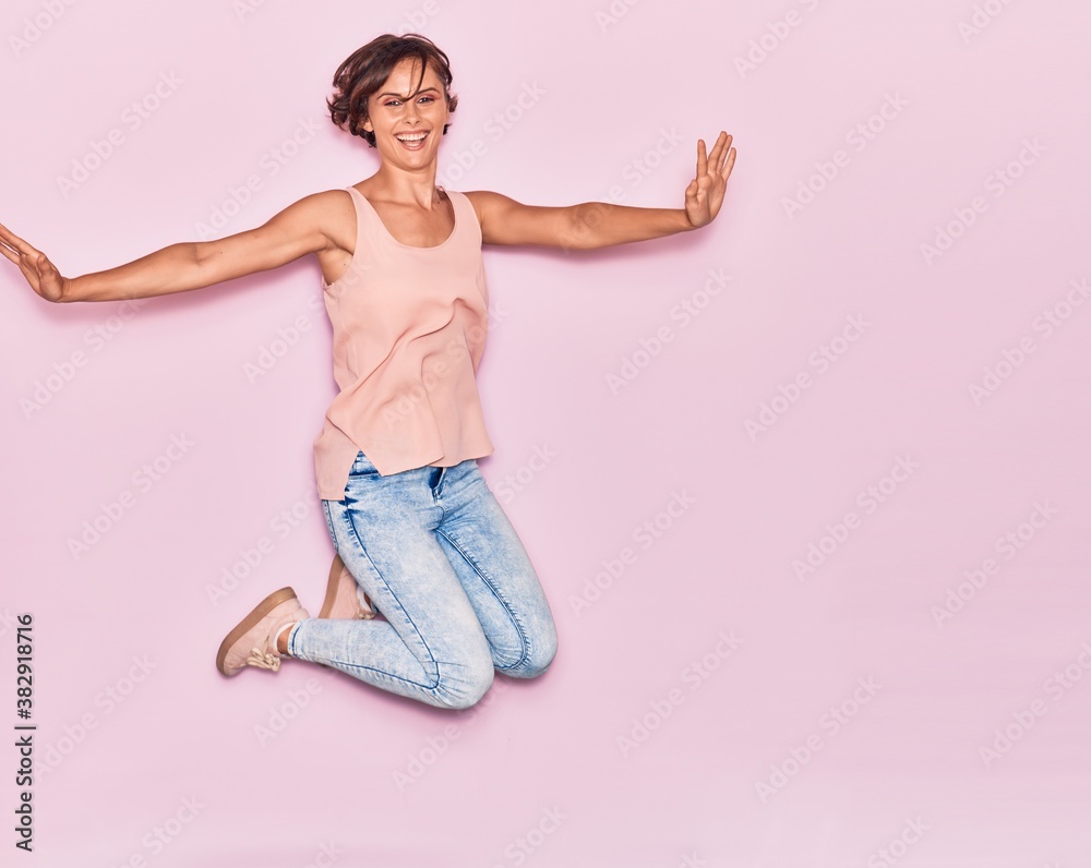 Young beautiful woman wearing casual clothes smiling happy. Jumping with smile on face and arms opened over isolated pink background. Young beautiful woman wearing casual clothes smiling happy. Jumping with smile on face and arms opened over isolated pink background.