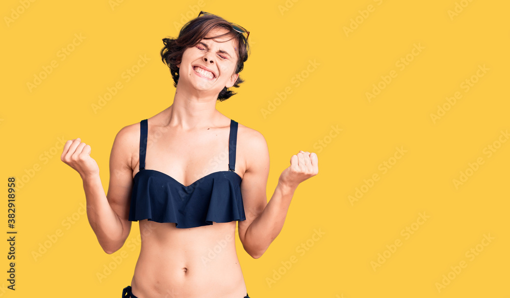 Beautiful young woman with short hair wearing bikini very happy and excited doing winner gesture with arms raised, smiling and screaming for success. celebration concept. Beautiful young woman with short hair wearing bikini very happy and excited doing winner gesture with arms raised, smiling and screaming for success. celebration concept.