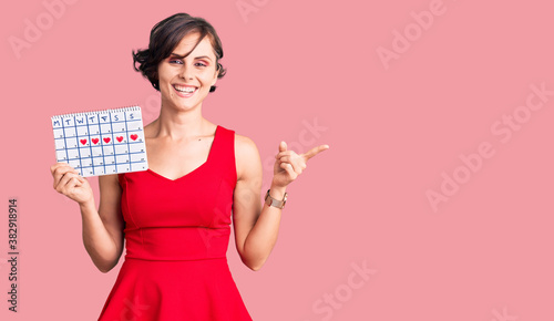 Beautiful young woman with short hair holding heart calendar smiling happy pointing with hand and finger to the side