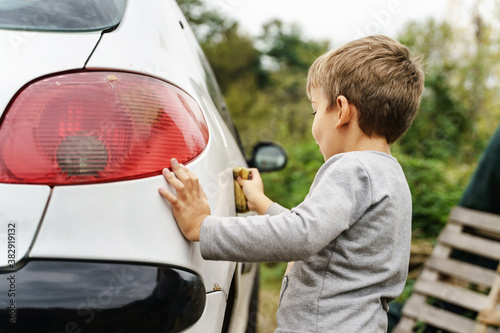 Small caucasian boy using sponge to wash the car in day outdoor - little child cleaning automobile side view - kid rubbing side of the white vehicle