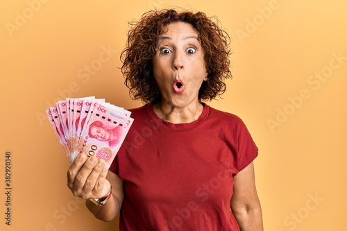 Beautiful middle age mature woman holding 100 yuan chinese banknotes scared and amazed with open mouth for surprise, disbelief face