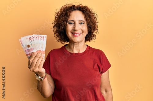 Beautiful middle age mature woman holding 10 colombian pesos banknotes looking positive and happy standing and smiling with a confident smile showing teeth