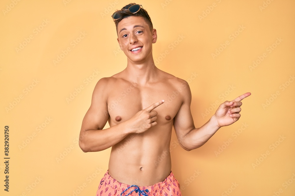 Young hispanic boy wearing swimwear shirtless smiling and looking at the camera pointing with two hands and fingers to the side.