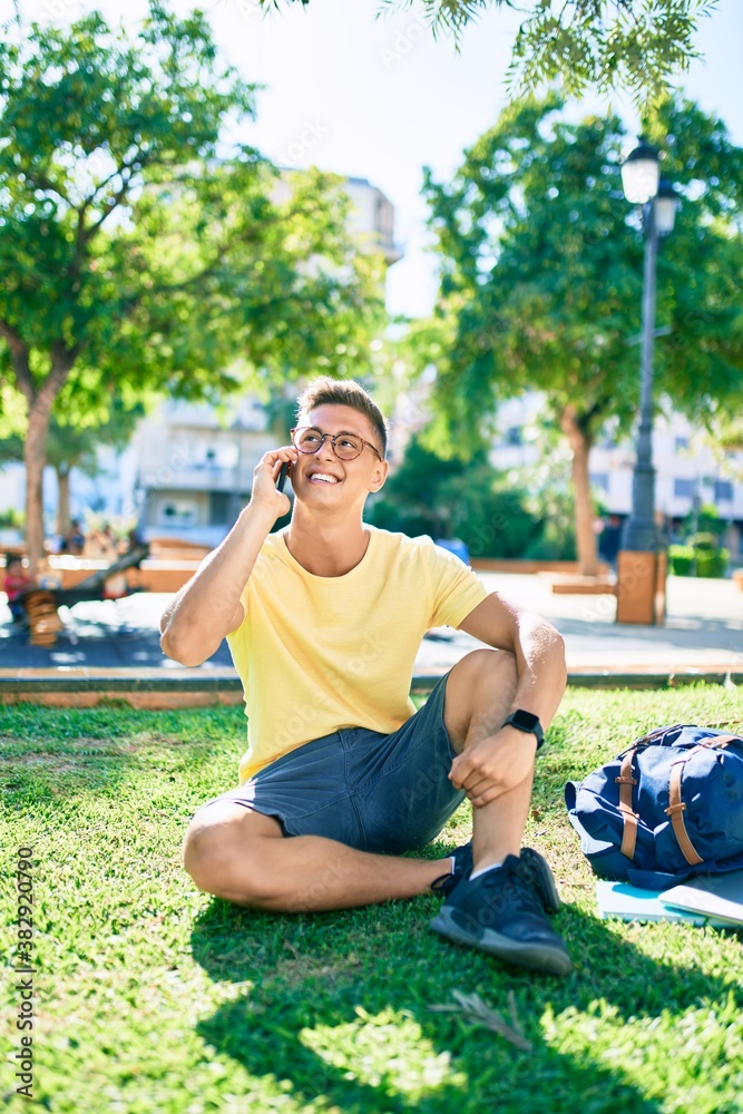 Fototapeta premium Young hispanic student smiling happy using smartphone sitting on the grass at the university campus