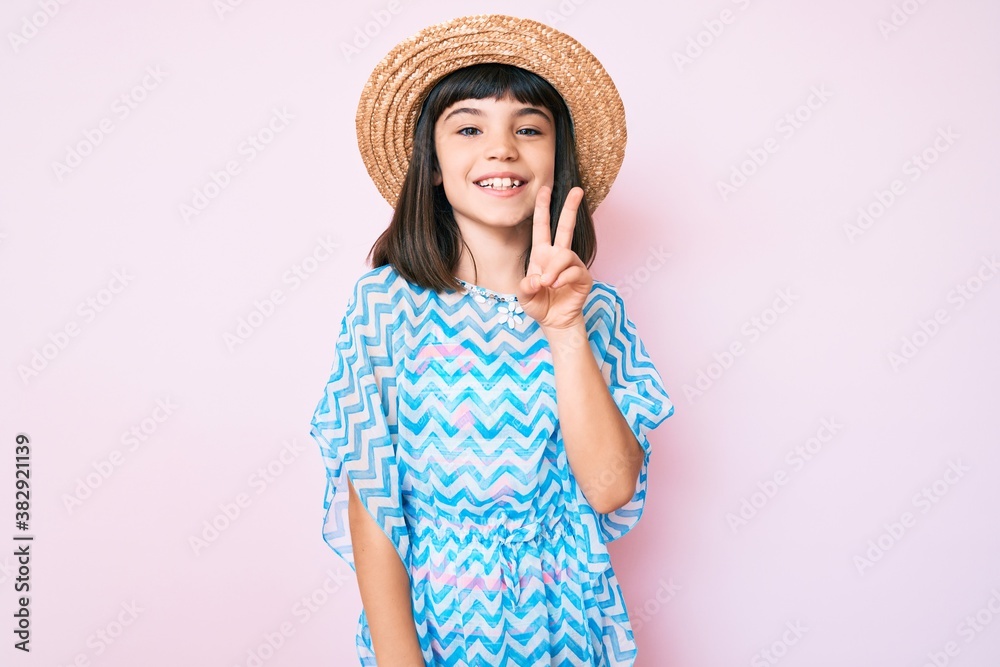 Young little girl with bang wearing summer dress and hat smiling with happy face winking at the camera doing victory sign. number two.