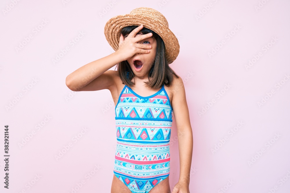 Young little girl with bang wearing swimsuit and summer hat peeking in shock covering face and eyes with hand, looking through fingers afraid Young little girl with bang wearing swimsuit and summer hat peeking in shock covering face and eyes with hand, looking through fingers afraid