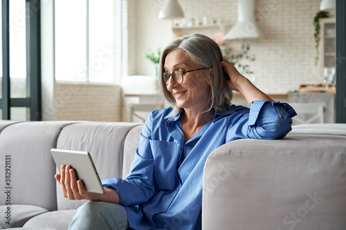 Happy 60s older mature middle aged adult woman holding digital tablet computer conference calling by social distance virtual family online chat meeting or watching video sitting on couch at home.