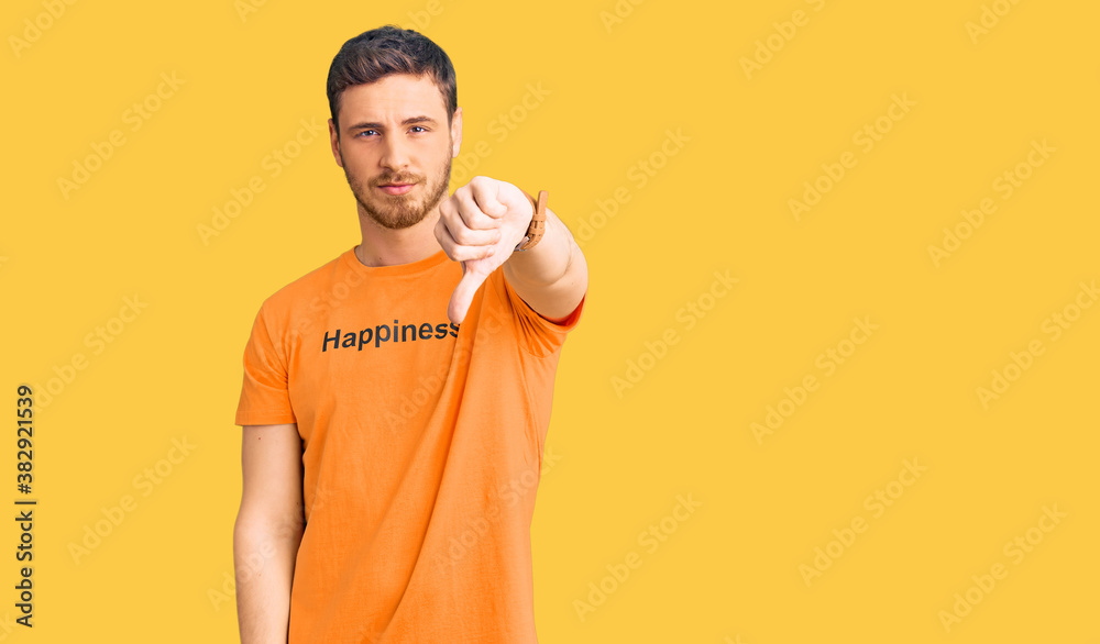 Handsome young man with bear wearing tshirt with happiness word message looking unhappy and angry showing rejection and negative with thumbs down gesture. bad expression. Handsome young man with bear wearing tshirt with happiness word message looking unhappy and angry showing rejection and negative with thumbs down gesture. bad expression.