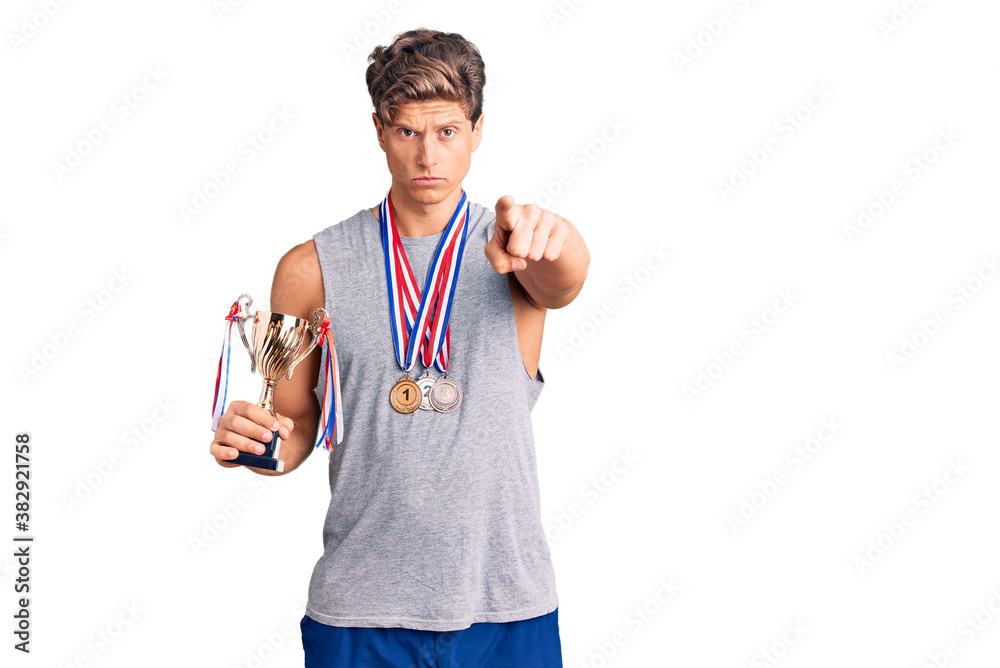 Young handsome man holding champion trophy wearing medals pointing with finger to the camera and to you, confident gesture looking serious Young handsome man holding champion trophy wearing medals pointing with finger to the camera and to you, confident gesture looking serious